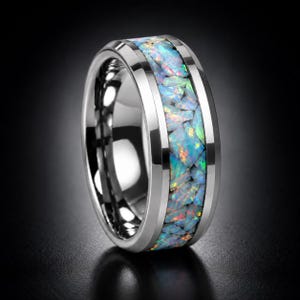 May include: A polished silver-toned metal ring featuring a wide center inlay composed of crushed iridescent opal fragments. The inlay displays a mosaic of light blue, green, and orange flecks that catch the light against the smooth, reflective metallic band.