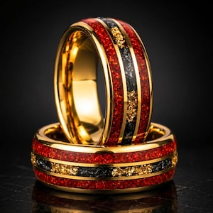 May include: Two gold-toned rings featuring a central inlay of crushed red material and black fragments with gold-colored flecks. The rings have a polished gold finish on the edges and a smooth interior surface, showcasing a layered design.