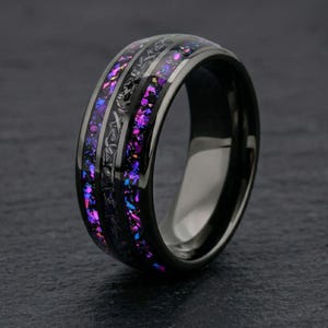 Könnte beinhalten: Ein schwarzer Ring mit einem polierten dunklen Band und einer zentralen Einlage aus zerkleinerten, irisierenden violetten und blauen Flocken. Die Einlage ist durch eine dünne, texturierte schwarze Linie unterteilt, was einen galaxieartigen Look erzeugt.