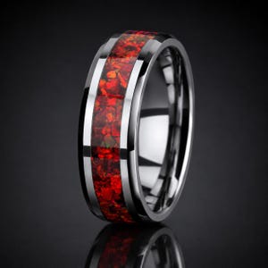 Puede incluir: Un anillo de tono plateado pulido con una incrustación central ancha de fragmentos de piedra roja triturada. La banda tiene bordes biselados y un acabado metálico liso y reflectante que contrasta con la incrustación roja texturizada.