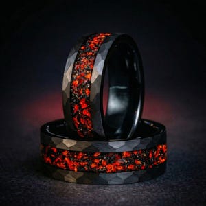 Op de afbeelding: Twee zwarte ringen met een gehamerde afwerking en een centrale inleg met een patroon van rode en zwarte steenslag. De ringen hebben een gladde binnenkant en een getextureerde buitenband, wat een contrast vormt tussen het donkere metaal en rood.