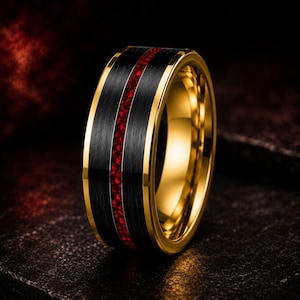 Puede incluir: Un anillo que presenta una banda central negra cepillada con una fina incrustación de color rojo vibrante que recorre horizontalmente el centro. Los bordes y el interior de la banda tienen un acabado en tono dorado pulido.