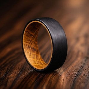 May include: A ring featuring a matte black outer band with a visible brushed texture and a smooth, light brown wood grain interior lining. The band has a rounded profile and a wide, solid construction, showcasing a contrast between the dark exterior and the natural, patterned wooden inner surface.