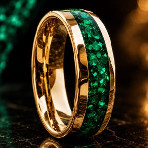 May include: A polished gold-toned ring featuring a central inlay of crushed green stone fragments. The band has a smooth, reflective metallic finish on both sides of the textured, vibrant green inlay, creating a distinct contrast in surface appearance.