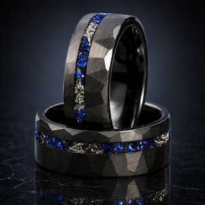 May include: Two dark gray rings with a faceted, hammered texture. Each band features a central inlay containing a mix of small, vibrant blue gemstones and textured, metallic silver-toned fragments running horizontally around the entire circumference.