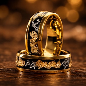 Op de afbeelding: Een paar goudkleurige ringen met een zwarte inlegband in het midden. De inleg is versierd met verspreide, onregelmatige goud- en zilverkleurige metaalvlokken. De ringen hebben gepolijste gouden randen en een glad, reflecterend binnenoppervlak.