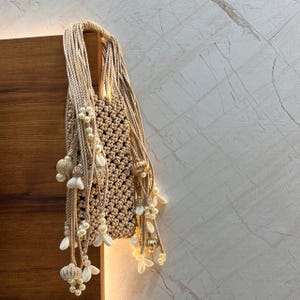 Puede incluir: Un bolso tote de macramé beige con un patrón tejido. La bolsa está adornada con perlas blancas y adornos en forma de flor. El asa está hecha de cuerda trenzada. La bolsa cuelga contra una superficie de madera y una pared de mármol.