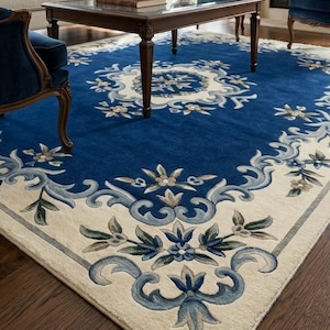 May include: A large rectangular area rug featuring a deep blue center with a central cream-colored floral medallion. The rug has a wide cream border decorated with intricate blue and light blue scrolling floral and leaf patterns throughout.
