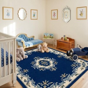 May include: A large rectangular area rug featuring a deep blue center with a cream-colored floral border and intricate scrollwork patterns. The rug is positioned in a room with a light-colored wooden floor, a white crib, a blue velvet chaise, and toys.