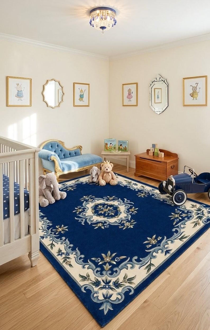 May include: A large rectangular area rug featuring a deep blue center with a cream-colored floral border and intricate scrollwork patterns. The rug is positioned in a room with a light-colored wooden floor, a white crib, a blue velvet chaise, and toys.