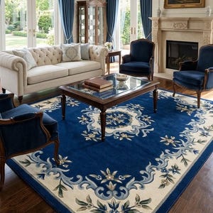 May include: A large rectangular area rug featuring a deep blue center and a wide cream-colored border. The rug is decorated with intricate floral and scrolling vine patterns in shades of light blue, sage green, and white throughout the design.