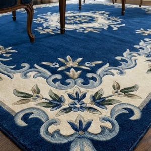 May include: A large rectangular area rug featuring a deep blue field with a cream-colored border. The design includes intricate floral motifs and scrolling acanthus leaf patterns in shades of light blue, sage green, and cream throughout the border.