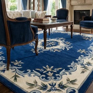 May include: A large rectangular area rug featuring a deep blue field with a cream-colored border. The design includes intricate floral motifs and scrolling acanthus leaf patterns in shades of light blue, grey, and white, creating a symmetrical border.