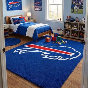 Może przedstawiać: Prostokątny niebieski dywan z dużym białym i czerwonym logo Buffalo Bills pośrodku. Dywan leży na drewnianej podłodze w sypialni urządzonej z pościelą w barwach drużyny, plakatami na ścianach i regałem wypełnionym przedmiotami o tematyce sportowej.