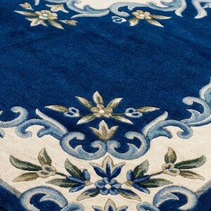 May include: A rectangular area rug featuring a deep blue background with a central tan medallion surrounded by cream-colored borders. The design includes intricate floral motifs and scrolling leaf patterns in shades of light blue, sage green, and white.