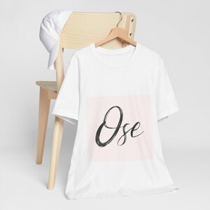 Peut inclure: T-shirt blanc avec le mot "Ose" en script noir sur un carré rose clair. Le t-shirt est suspendu à un cintre en bois fixé à une chaise en bois. Une casquette de baseball blanche repose sur la chaise.