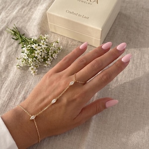 Puede incluir: Una cadena de mano dorada con tres gemas en forma de marquesa. Esta delicada joya conecta con un anillo y una pulsera. La mano descansa sobre una superficie de color neutro, con un pequeño ramo de flores blancas y una caja de joyas en el fondo.