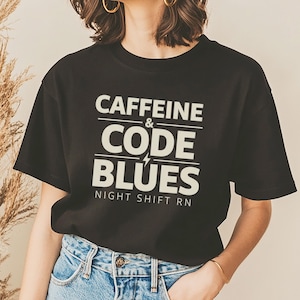 Puede incluir: Camiseta negra con el texto blanco "CAFFEINE & CODE BLUES NIGHT SHIFT RN" y un rayo. La camiseta se combina con unos vaqueros azules.