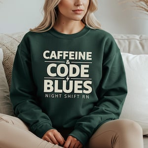 Puede incluir: Sudadera verde oscuro con el texto blanco "CAFFEINE & CODE BLUES" y un rayo. Debajo está escrito "NIGHT SHIFT RN". La sudadera es de cuello redondo.