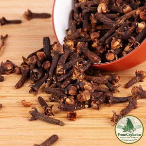 May include: A collection of dried whole cloves spilling out of a red bowl. The dark brown, nail-shaped spices feature a textured stem and a rounded, four-petaled flower bud at the top. A circular logo with the text FromCeylonco is visible.