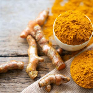 Puede incluir: Primer plano de raíces y polvo de cúrcuma. La imagen muestra varios rizomas de cúrcuma frescos con un tono naranja brillante, junto con un cuenco blanco y una cuchara de madera llenos de vibrante polvo de cúrcuma amarillo. La escena está ambientada sobre una superficie de madera rústica.