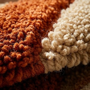 May include: Close-up of a textured rug with a color block design. The rug features a combination of warm orange and cream-colored sections. The looped pile creates a soft, inviting texture, suggesting comfort and warmth. The rug's design is simple yet visually appealing.