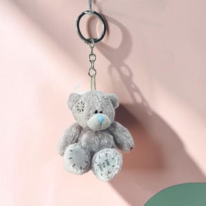 May include: A small, gray teddy bear keychain with a silver keyring. The bear has a blue nose and stitched details. The bear is hanging against a pink and green background.