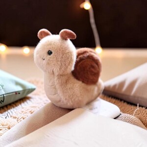 May include: A soft toy snail with a light brown body and a dark brown shell. The snail has small, rounded ears and is placed on a cream-coloured surface. A light green cushion with black embroidered designs is in the background.