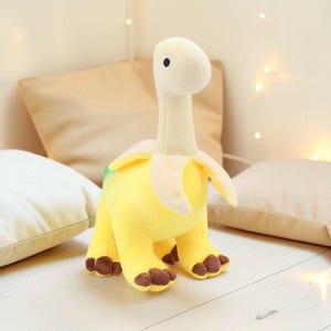 Puede incluir: Un peluche de dinosaurio con diseño de cáscara de plátano. El dinosaurio es amarillo con cuello y cabeza color crema y pies marrones. Está rodeado de almohadas blancas y beige, con luces de cadena en el fondo.