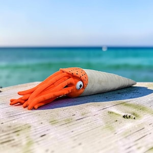Peut inclure: Une peluche en forme de fossile d'ammonite avec une tête orange vif dotée de multiples tentacules et d'un œil blanc, fixée à une longue coquille beige effilée à la texture lisse et au motif tacheté près de la tête.