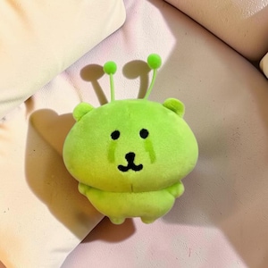 May include: A bright lime green plush toy shaped like a rounded creature with two thin antennae topped with small spheres, featuring embroidered black eyes, a small nose, and a mouth, with two small arms and legs extending from the body.