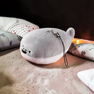 May include: A plush gray seal toy with black embroidered facial features and a black lanyard. Several white decorative pillows with black and red patterns surround the seal toy. The pillows have various floral and geometric designs.