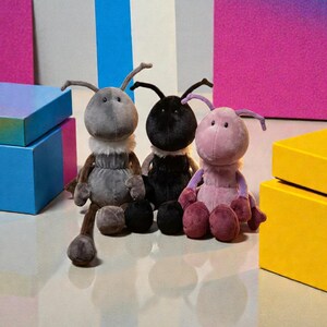 Könnte beinhalten: Drei Plüsch-Ameisen-Spielzeuge in Grau, Schwarz und Rosa, jeweils mit einem flauschigen weißen Kragen und langen Beinen. Die Spielzeuge stehen vor einem Hintergrund aus bunten Boxen und einem Farbverlauf, was eine verspielte Szene erzeugt.