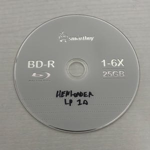 Puede incluir: Un disco BD-R plateado con el logotipo "smartbuy" y el texto "BD-R", "1-6X" y "25GB". El texto escrito a mano en el disco dice "HENLOADER LP 1A". El disco tiene una superficie reflectante.