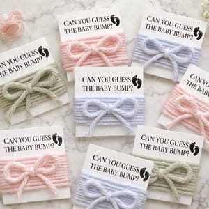 Puede incluir: Tarjetas con el texto "CAN YOU GUESS THE BABY BUMP?" están envueltas con hilo en colores pastel, incluyendo rosa, azul y verde. Cada tarjeta tiene un lazo hecho del mismo hilo. Un chupete y flores también están en la imagen.