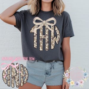 May include: Dark gray t-shirt featuring a monogram design with a camouflage pattern and a light pink bow. The monogram includes the letters THW. The text "AVAILABLE WITH PINK BOW" is visible.