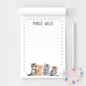 May include: A white notepad with the name "MARCIE WILLIS" printed at the top. The notepad features a dotted border and a watercolor illustration of four kittens in various colors at the bottom. A pencil rests on the right.
