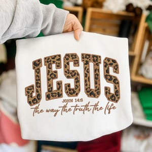 May include: Cream-coloured sweatshirt featuring the word "JESUS" in leopard print lettering. Beneath, the text reads "JOHN 14:6" and "the way the truth the life" in a brown script. The sweatshirt is held by a person.