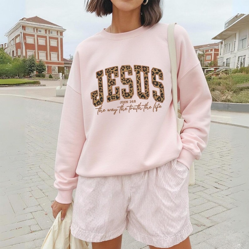 May include: A pale pink sweatshirt featuring the word "JESUS" in a leopard print pattern. The text "JOHN 14:6 The way the truth the life" is below. The person is also wearing pink shorts.