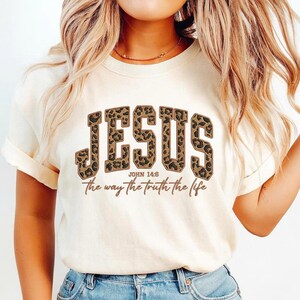 May include: A cream-coloured t-shirt featuring the word "JESUS" in large leopard print lettering. Underneath, the text reads "JOHN 14:6" and the phrase "the way the truth the life" in a brown script font.