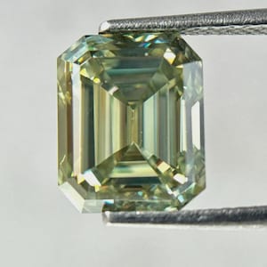 May include: An emerald-cut gemstone with a pale greenish-yellow hue and high clarity. The stone features sharp, linear facets that create a geometric pattern of light reflections, held securely by metal tweezers at the top and bottom edges.