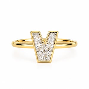 Op de afbeelding: Een gouden ring met een opvallende letter V als middelpunt. De letter V is ingelegd met meerdere kleine, heldere, gefacetteerde stenen die het licht vangen. De ringband is van glad, gepolijst goud, wat zorgt voor een strak en reflecterend oppervlak.