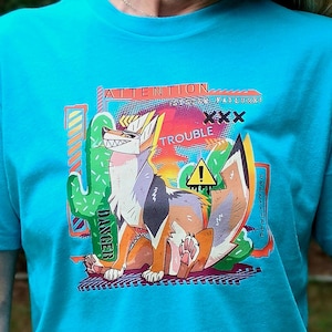 May include: A bright turquoise short-sleeve t-shirt featuring a graphic print of a stylized canine character. The design includes a cactus, a yellow warning sign, and the text "ATTENTION", "SYSTEM FAILURE", and "TROUBLE".