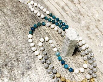 Könnte beinhalten: Eine mehrsträngige Mala-Halskette mit blauen, weißen und braunen Perlen. Die Halskette besteht aus natürlichen Steinen und ist auf einer goldenen Schnur aufgefädelt. Die Halskette wird auf einer Holzoberfläche gezeigt.