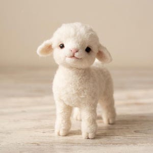 Pode incluir: Figura de cordeiro branco artesanal, com textura de lã fofa. O cordeiro tem olhos pequenos e escuros, nariz rosa e uma expressão gentil. O cordeiro está em pé sobre uma superfície de madeira clara.