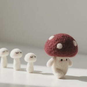 Puede incluir: Una caprichosa figura de seta de fieltro con un gorro burdeos y lunares blancos, caminando delante de tres setas blancas más pequeñas. La seta tiene una cara sonriente y brazos pequeños. Las setas están dispuestas sobre una superficie blanca.