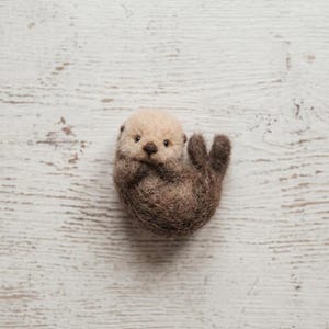May include: A needle-felted sea otter figurine with a light beige head and a dark brown body. The otter is curled into a rounded shape with its small paws tucked near its face, featuring dark bead eyes and a small dark nose made of felted wool.