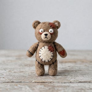 May include: A handcrafted brown teddy bear with stitched patches and a heart-shaped patch. The bear has wide, surprised eyes and a stitched mouth. The bear is standing on a wooden surface.