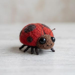 May include: A needle-felted ladybird figurine crafted from wool. The body is bright red with dark brown circular spots. It features a brown head section with two thin antennae and two large, glossy black bead eyes. It has six small dark brown legs.
