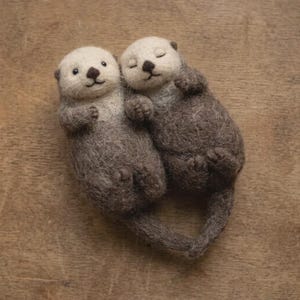 May include: Two needle-felted otter figurines with textured dark brown bodies and lighter cream-coloured faces. The otters are positioned side-by-side, with one otter having its eyes open and the other with its eyes closed, holding their tails together.